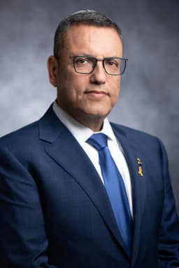 צילום תדמית רשמי של משה ליאון, ראש עיריית ירושלים, בחליפה כהה ועניבה כחולה על רקע אפור ממוזג — תאורת סטודיו רכה ומבט ישיר למצלמה