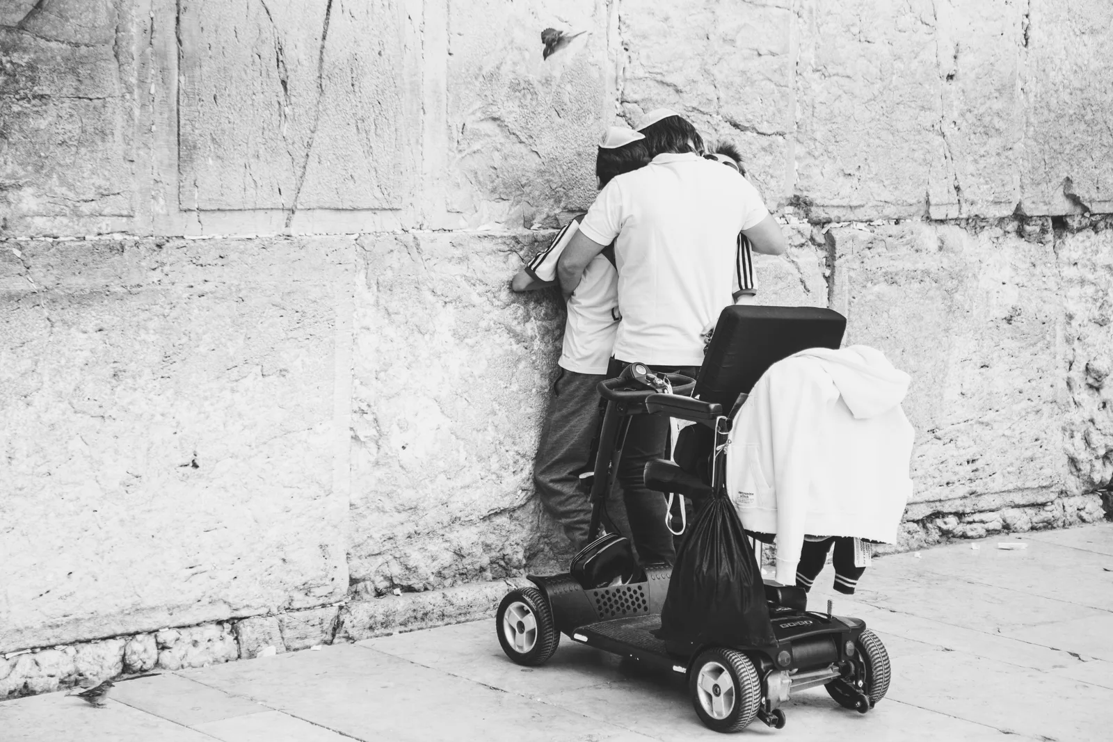 אב מרים את בנו לנשק את אבני הכותל המערבי בזמן בר מצווה, ציפור מרחפת מעל — צילום רחוב של שרון גבאי מתוך הספר ירושלמים
